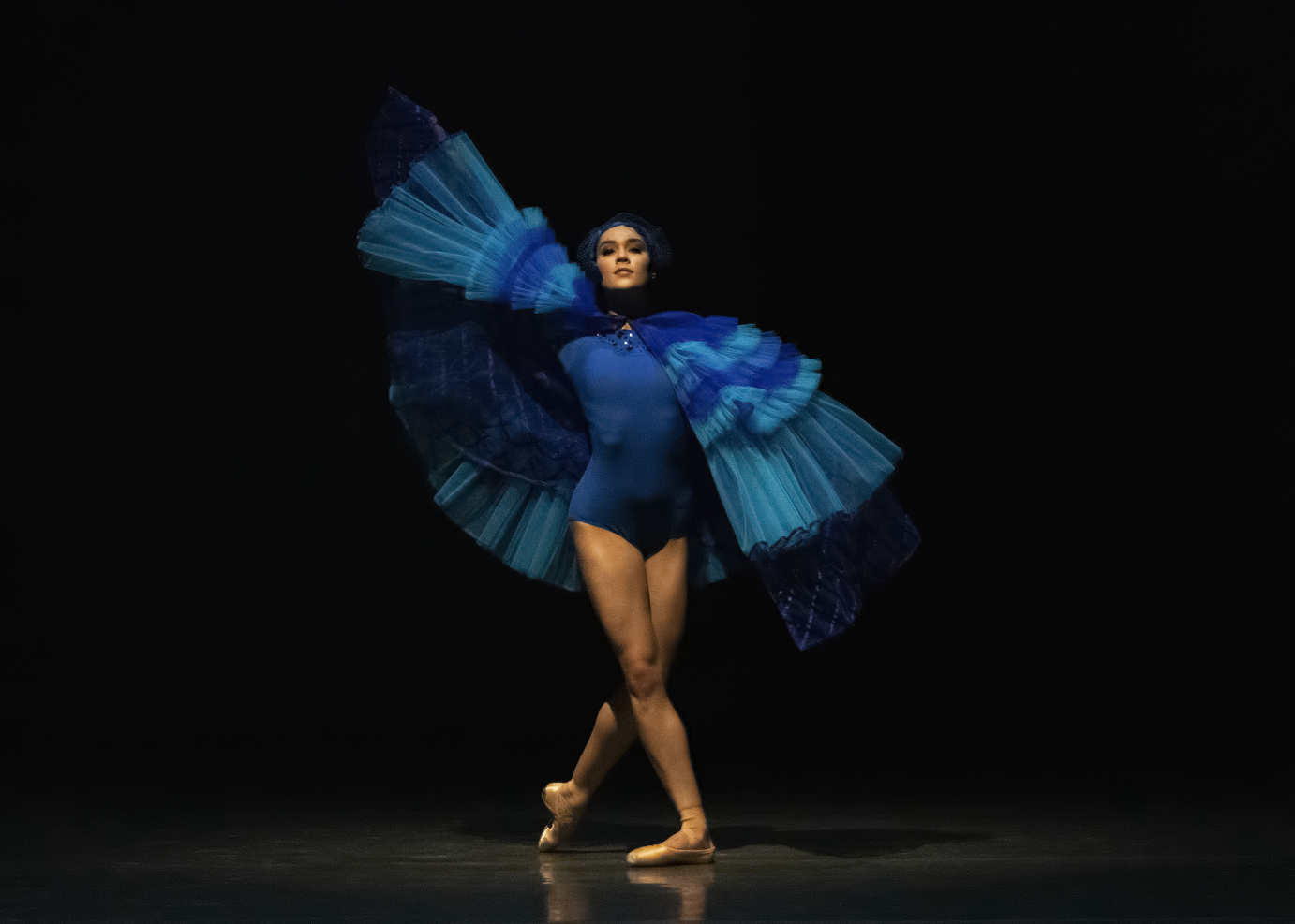 A woman in a blue leotard with tulle wings stands in B-plus, facing the camera straight on.